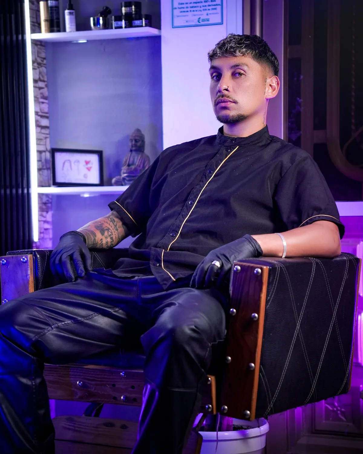Grey Gamboa sentado en su silla de barbero, con camisa negra de ribete dorado y guantes negros, bajo luz violeta